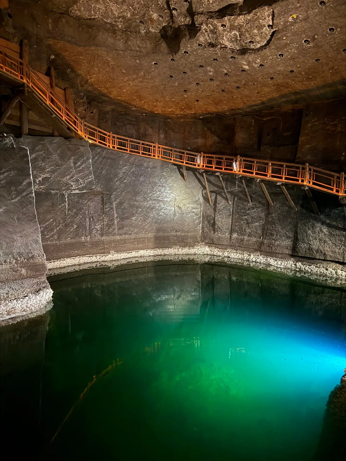 The underground salt lake in the Wieliczka Salt Mine with turquoise-illuminated water and a wooden jetty for tourists along the raw walls of the excavation.
